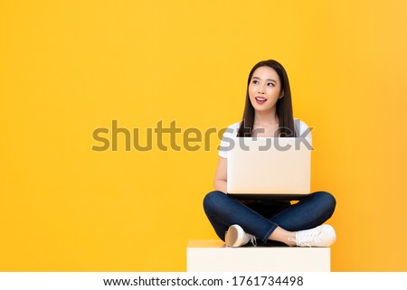 Happy attractive young Asian woman with laptop computer sitting cross legged and looking at empty space on yellow studio background