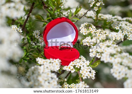 A gold wedding ring in a red heart-shaped gift box lies among many small white flowers: a wedding concept, a place for text