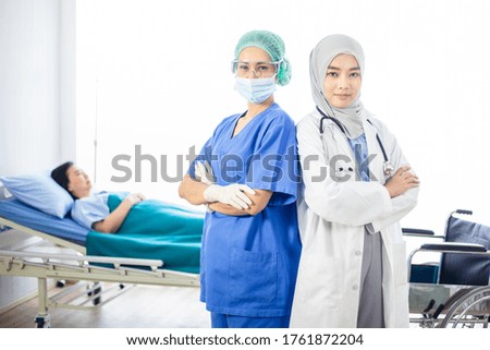 Medical staff looking at the camera and blurred senior patient on background. Muslim doctor and masked Asian nurse visiting senior woman and having discussion in a hospital. 