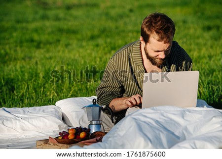 Man sitting in bed outdoors in grassfield in the middle of nowhere, keen working on his laptop under a gentle spring sun.