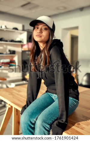 Cheerful female worker looking at camera while posing in custom apparel, baseball cap and hoodie. Focus on a girl. Young woman working at custom T-shirt, clothing printing company. Vertical shot