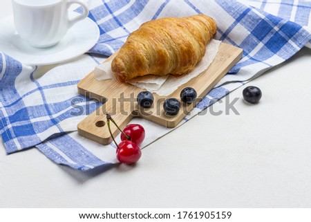 Croissant on white and blue checkered napkin. Cup of coffee cherry and blueberries on table. Top view, White background
