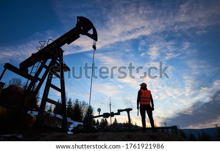 Horizontal snapshot of an oilman standing with his back to the camera, holding a pipe wrench and looking at the oil pump jack, beautiful silhouettes against blue sky, concept of oil and gas industry