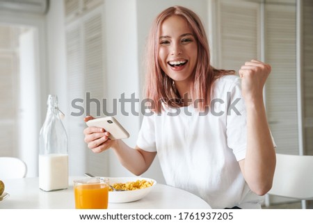 Photo of young excited woman making winner gesture while playing online game on cellphone in white kitchen