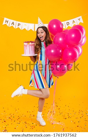 Image of excited cute woman in party cone posing with pink balloons and birthday torte isolated over yellow background