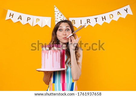Image of excited woman in party cone showing birthday torte and blowing whistle isolated over yellow background