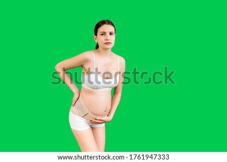 Portrait of pregnant woman in underwear with bandage against backache at green background with copy space. Mother is suffering from pain in the back. Orthopedic abdominal support belt concept.