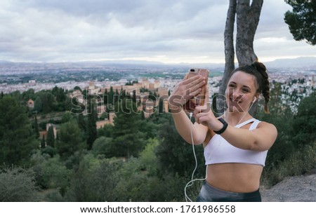 Smiling pretty young girl taking selfies in a viewpoint with the Alhambra in the background after practicing yoga. Woman enjoying in nature wearing sportswear above the city listening to music.