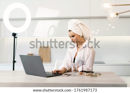 Cute young attractive woman with a towel on her head doing morning skin care procedures. Girl is watching news on a laptop after a shower. Blogger woman is ready to stream about beauty.