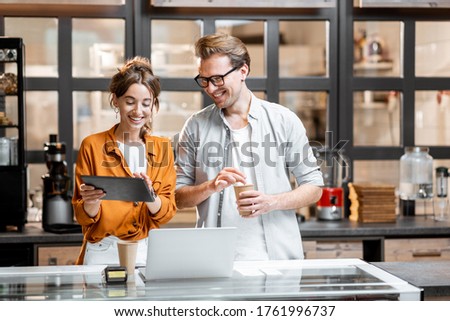 Two young managers or shop owners having some discussion while standing with a digital tablet at the counter of the shop or cafe. Small business management concept Royalty-Free Stock Photo #1761996737