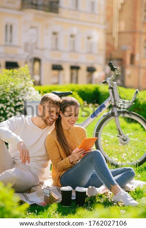 Great mood. Interested satisfied woman with tablet and man sitting close together on grass on plaid, smiling looking at the screen