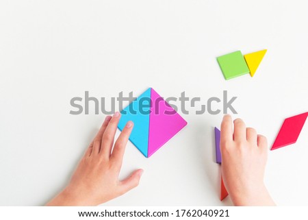 Tangram colored geometric puzzle pieces with childs hands moving pieces on white table, top view. 