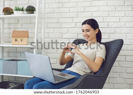 Young brunette chatting online, making hearth sign. Side view of video call communication.
