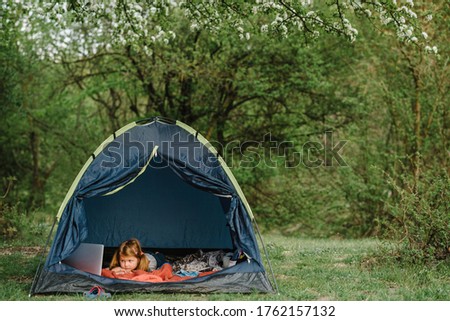 Children tourism. Child using laptop in the tent at the campsite. Girl watching cartoon on gadget. Kid girl in a campaign in a tent. Family summer vacation in nature.