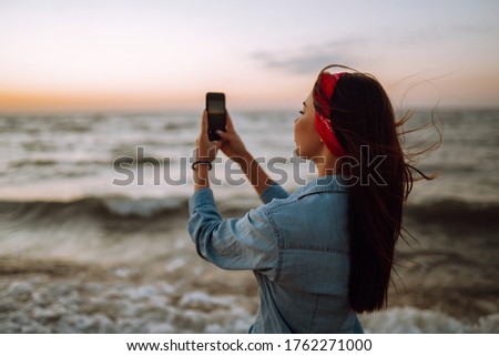 Young girl sitting on the beach photographs the sea at sunset. Woman on vacation using mobile phone to take photo of sea waves.