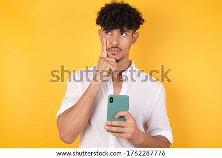 Image of a thinking dreaming young handsome man posing isolated over yellow wall background using mobile phone and holding hand on face. Taking decisions and social media concept.