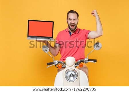 Joyful young bearded guy in summer clothes driving moped isolated on yellow background. Driving motorbike transportation concept. Hold laptop pc computer with blank empty screen, doing winner gesture