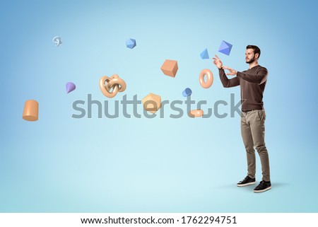 Handsome bearded young man in casual clothes standing and levitating lots of different geometric objects on light blue background. Thinking processes. Imagination and creativity. Royalty-Free Stock Photo #1762294751