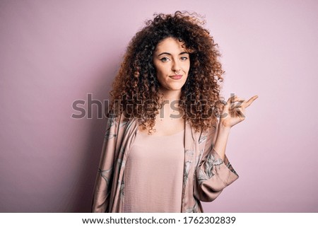 Young beautiful woman with curly hair and piercing wearing casual pink t-shirt with a big smile on face, pointing with hand and finger to the side looking at the camera.