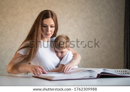A middle-aged Caucasian woman hugs a little son and shows a photo album. A little boy sits on his mothers lap and looks at old photographs. Pleasant memories. Close family relationships.