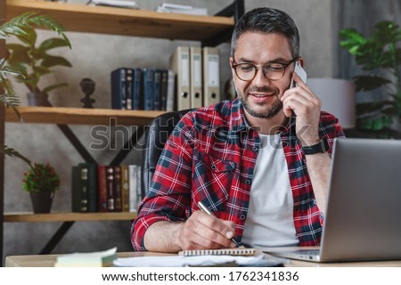 Smiling man writing notes while making phone call and using laptop at home Royalty-Free Stock Photo #1762341836