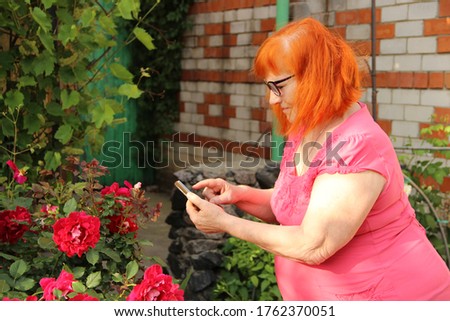 Fat old redheaded woman in a pink dress takes a picture of a rose on her phone.
