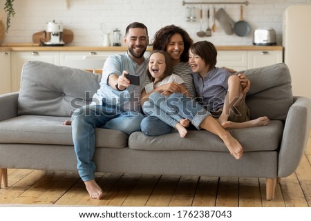 Smiling parents with little kids laughing using smartphone together sitting on couch at home. Happy father holding phone taking selfie with children. Family watching video having fun with cellphone.