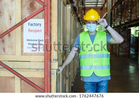 caucasian factory warehouse worker wearing face mask standing beside annoucement banner of factory reopen in front of factory warehouse during coronavirus covid-19 pandemic