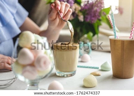 Woman is picking up a spoon and gently stirs the thick whipped mousse in a glass of cold coffee. Gentle atmosphere and sunny beautiful summer morning.