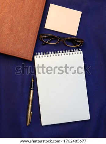 Notepad, pen and glasses on a dark blue fabric background. Space for your logo, layout.