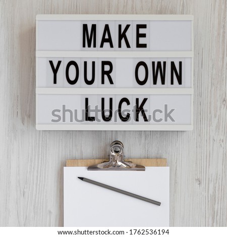 'Make your own luck' on a lightbox, clipboard with blank sheet of paper on a white wooden surface, top view. Flat lay, from above, overhead. 