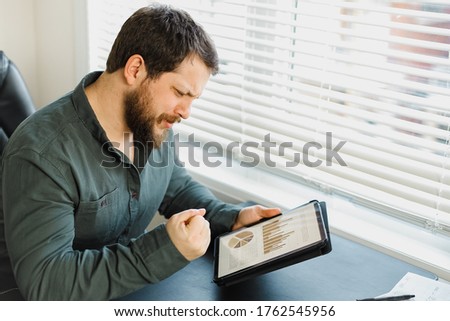 Male economist working with graphs and diagrams on tablet at office. Concept of professsional occupation and smart employee. Adult bearded man wearing shirt and surfing internet at cabinet.