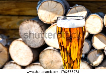 A full glass of fresh beer. Beer glass and timber. A GLASS WITH LIGHT BEER ON THE BACKGROUND OF wood in the countryside in the country. Beer glass and firewood. In the rays of sunlight. Close-up