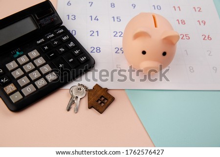 Day of buying or selling a house or payment for rent or loan. Piggy bank, wooden house keyring and calculator on white clean calendar