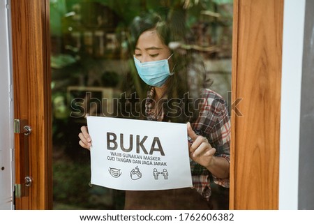 cafe open for business during new normal corona virus outbreak. woman cafe owner put sign in bahasa indonesia meaning : Open, put your mask on and keep your distance