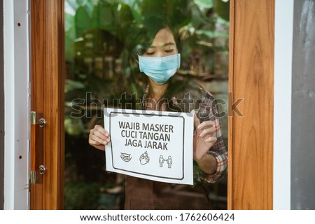 cafe open for business during new normal corona virus outbreak. woman cafe owner put sign in bahasa indonesia meaning : Open, put your mask on and keep your distance