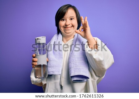 Young down syndrome fitness woman training at gym holding water bottle and sport towel doing ok sign with fingers, excellent symbol