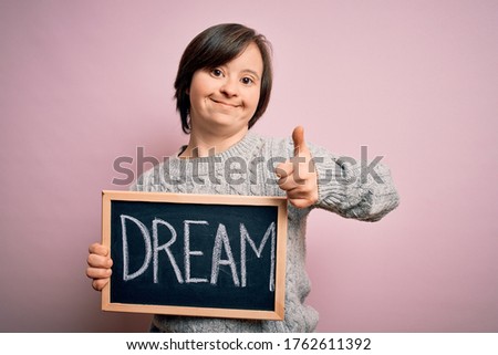 Young down syndrome woman holding blackboard with dream word as message of happiness happy with big smile doing ok sign, thumb up with fingers, excellent sign