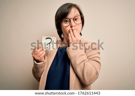 Young down syndrome business woman holding question mark paper as doubt symbol cover mouth with hand shocked with shame for mistake, expression of fear, scared in silence, secret concept