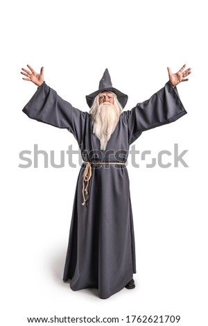 A stern grey-haired bearded wizard in a gray cassock and a cap is practicing sorcery and doing magic against a white background. Royalty-Free Stock Photo #1762621709