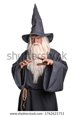 A stern grey-haired bearded wizard in a gray cassock and a cap is practicing sorcery and doing magic against a white insulating background. Royalty-Free Stock Photo #1762621751
