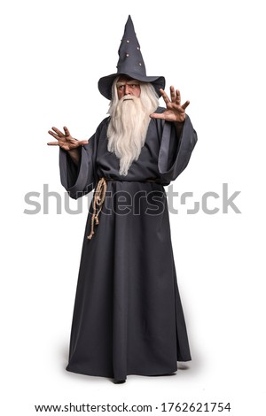 A stern grey-haired bearded wizard in a gray cassock and a cap is practicing sorcery and doing magic against a white insulating background. Royalty-Free Stock Photo #1762621754