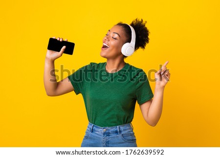 Excited emotional black woman singing her favorite song, using smartphone as microphone, yellow studio background Royalty-Free Stock Photo #1762639592