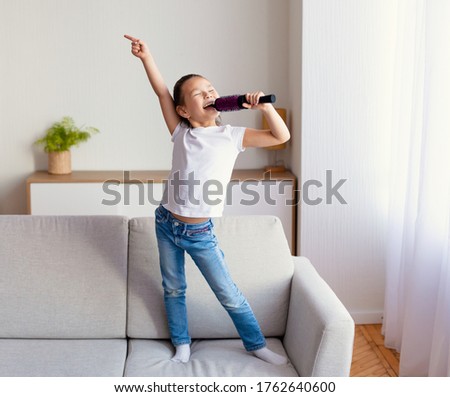 Little Singer. Adorable Girl Singing Song Holding Hairbrush Like Microphone Having Fun Standing On Sofa At Home Royalty-Free Stock Photo #1762640600