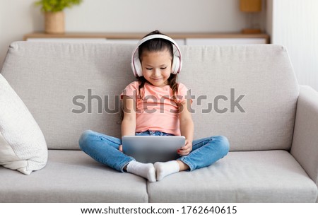 Asian Girl Using Tablet Computer Wearing Wireless Headphones Watching Cartoons Sitting On Couch At Home. Kid And Gadget Concept