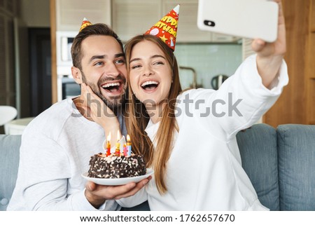 Cheerful attractive young couple celebrating birthday with cake, sitting on a couch at home, taking a selfie