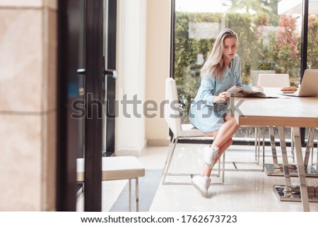Enthusiastic beautiful young woman housewife reads her favorite newspaper in kitchen while drinking tea. Young woman is up to date with world news. Modern interior,large Windows overlooking the garden