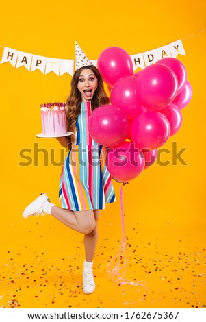 Image of excited cute woman in party cone posing with pink balloons and birthday torte isolated over yellow background