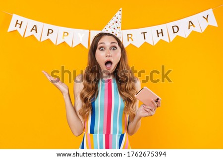 Image of shocked cute woman in party cone playing online game on cellphone isolated over yellow background