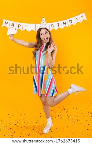 Image of joyful cute woman taking selfie on mobile phone and waving hand isolated over yellow background